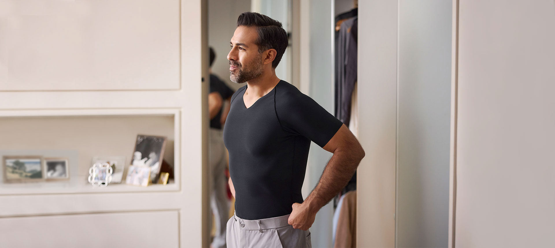 A picture of a man getting dressed. He is wearing a black compression shirt and gray pants, standing in a well lit bedroom.